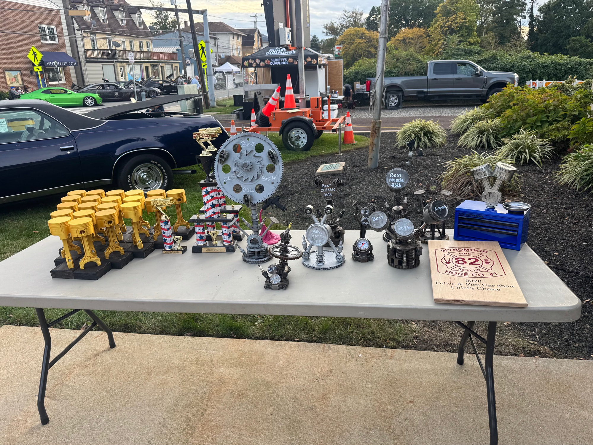 The handmade trophy table at the 2026 Wyndmoor Police and Fire Car Show, with cars and the vendor area behind it. Photo by Jim Semen.